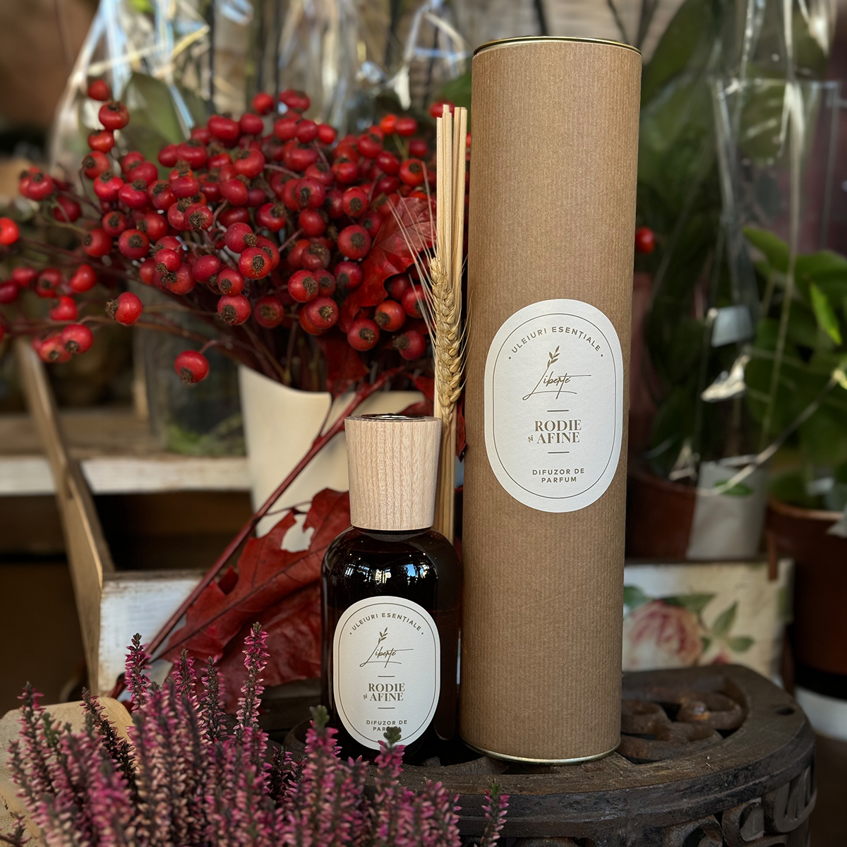 Fragrance gift set with a brown perfume bottle, wooden cap, and a tall diffuser tube on a rustic display with red berries and foliage as decor.